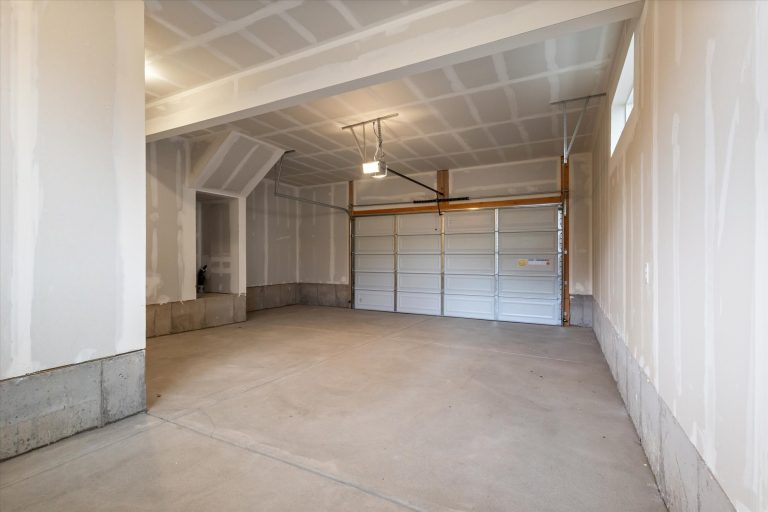 A clean, empty garage with unfinished drywall, a concrete floor, and a closed double garage door. Overhead lighting is on, and there is a small window near the ceiling on the right wall.