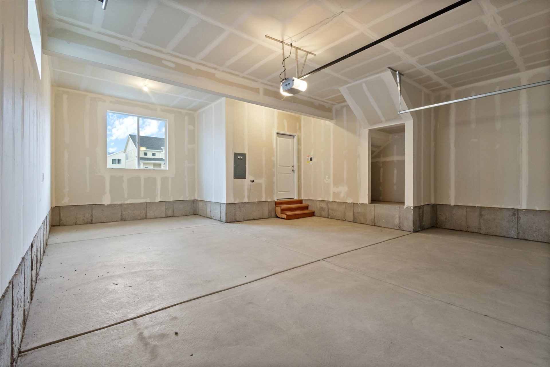 Unfinished two-car garage with bare drywall, concrete floor, a window, electrical panel, steps leading to a white door, and an open garage door track on the ceiling.