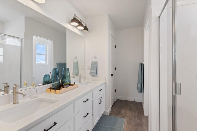 Modern bathroom with double sinks, white countertops, black hardware, decorative blue glass vases, large mirror, shower with glass door, and light wood flooring. Blue towels and a window add a fresh touch.