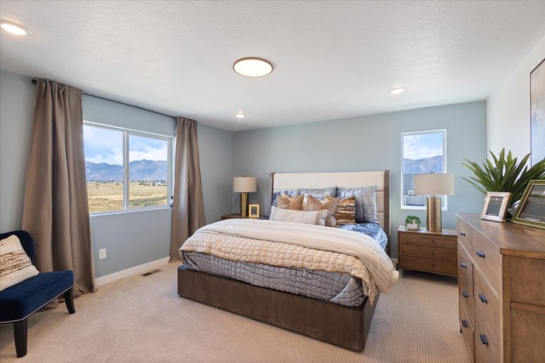 A modern bedroom with a large bed, gray and beige bedding, nightstands with lamps, two windows showing a mountain view, a dresser with decor, neutral walls, and beige carpet.