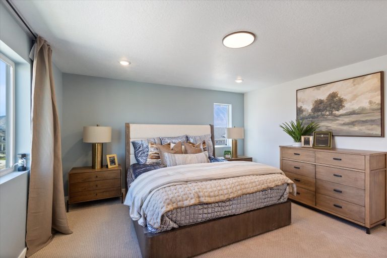 A modern bedroom with a large bed, wooden nightstands and dresser, beige carpet, neutral bedding, decorative pillows, framed artwork, potted plant, and two windows letting in natural light.