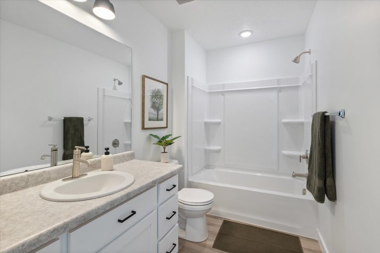 A modern bathroom with white walls, a vanity with a sink and faucet, a large mirror, a toilet, a bathtub with built-in shelves, green towels, a potted plant, wall art, and wood-look flooring.