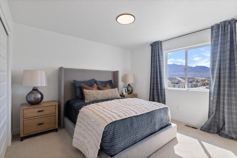 A bright bedroom with a gray upholstered bed, navy and beige bedding, two nightstands with lamps, plaid curtains, and a large window showing a mountain view.