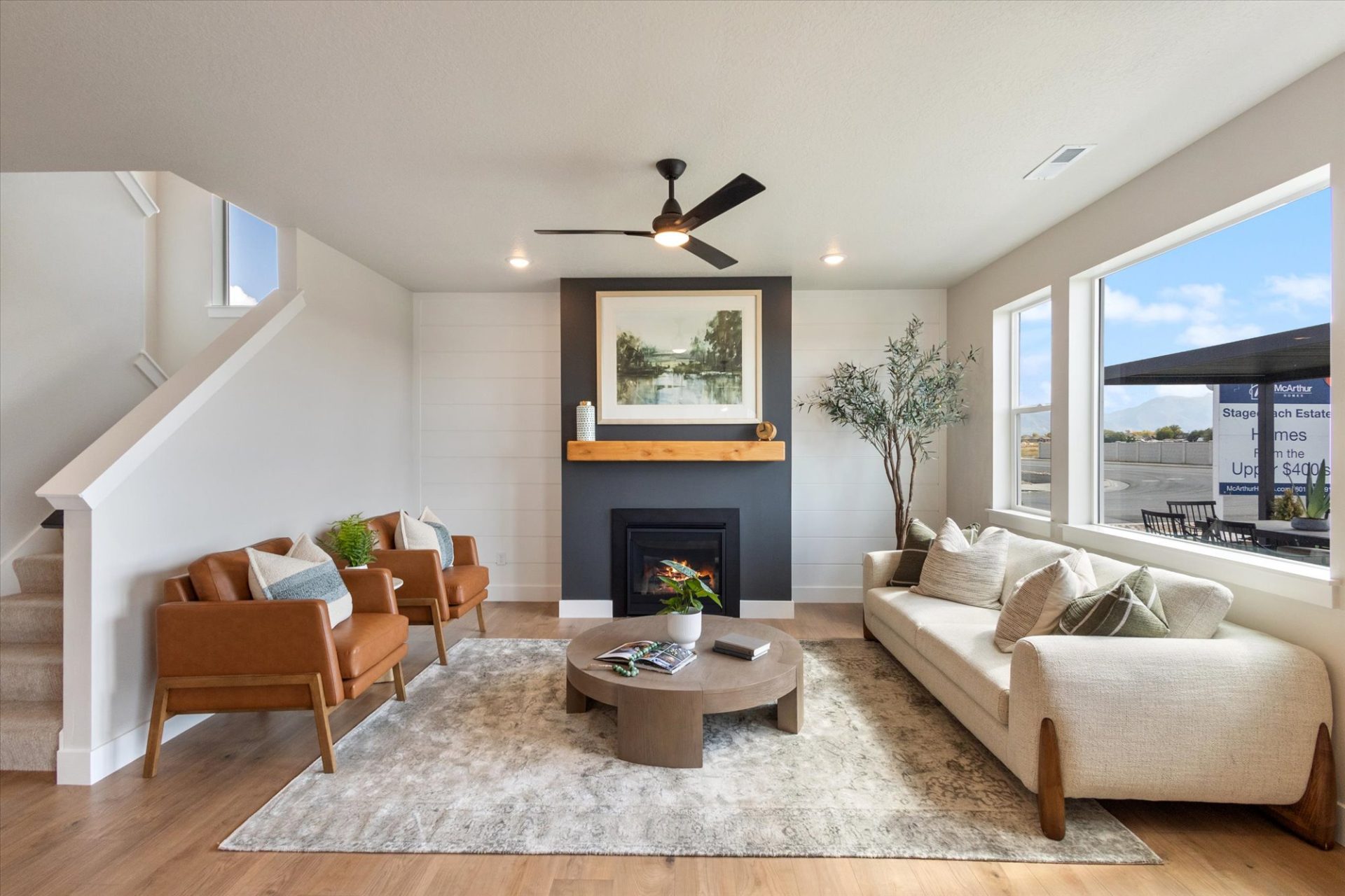 Open-concept living room with hardwood floors, dark shiplap fireplace, tan leather chairs, cream sofa with green pillows, and large windows.