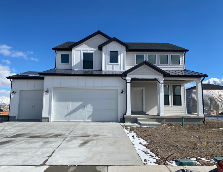 A newly built two-story house with white siding, black roof, and a three-car garage. Like many homes in Utah, it features a concrete driveway and a yard of bare dirt with patches of snow. Blue sky and mountains complete the view.
