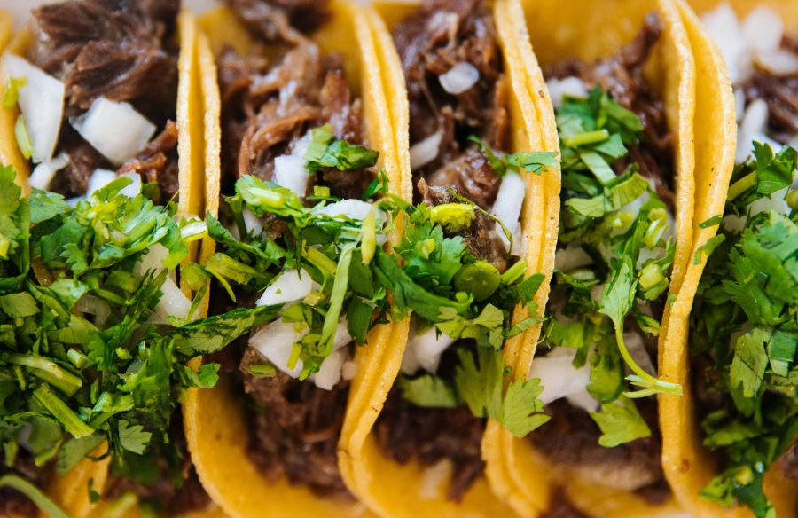 A row of tacos with onions and proteins, garnished with fresh greens and served on a long tray.