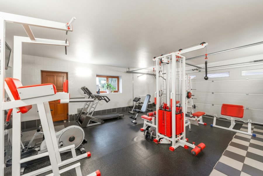 Modern home gym with various workout equipment including weight machines, treadmill, stationary bike, and strength training benches. Well-lit fitness room with red and white exercise machines and black rubber flooring.