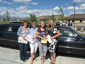 Four women stand smiling in front of a black limousine, each holding certificates. Behind them, homes in Utah, trees, and mountains complete the sunny outdoor scene.