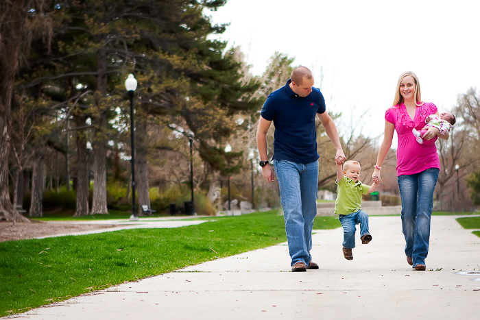 A man and woman walk outside on a path near homes in Utah, swinging a young boy between them. The woman holds a baby in her other arm. Trees and grass line the sidewalk, and all appear happy and smiling.