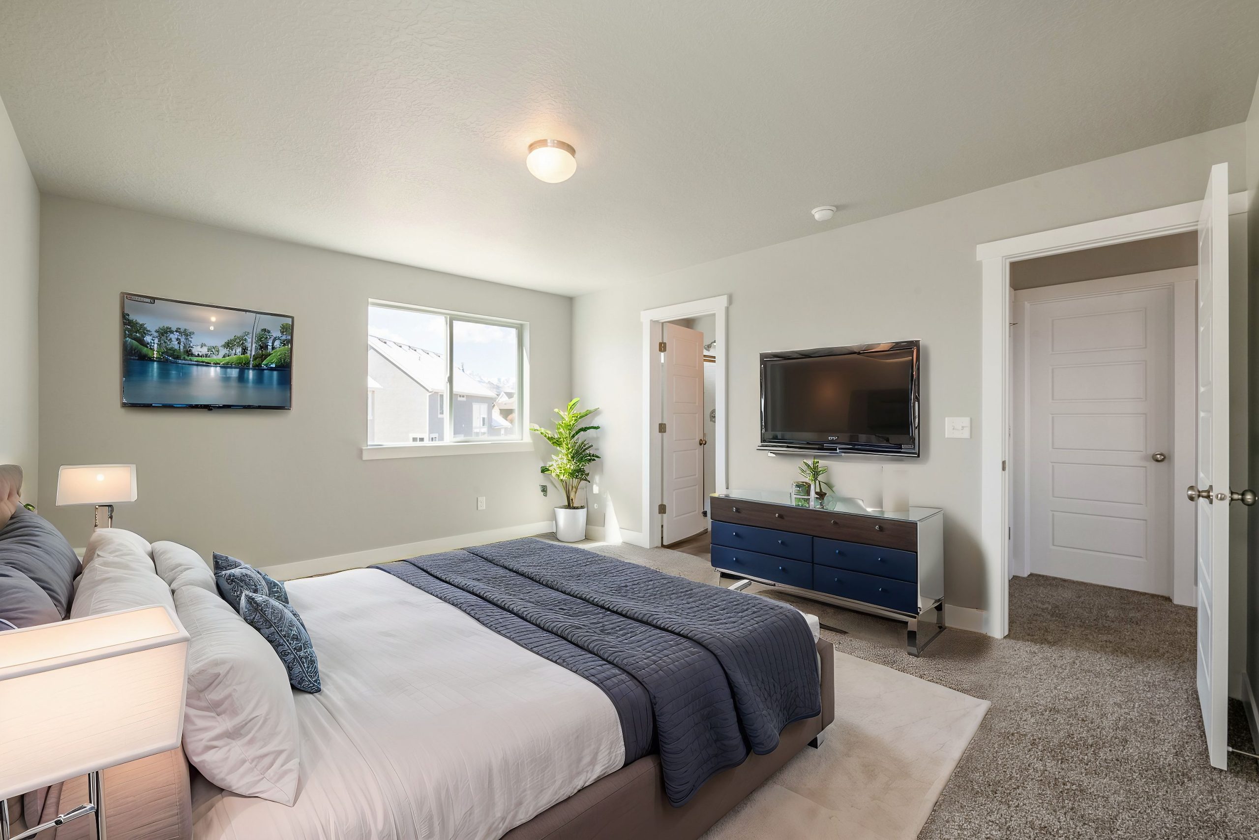 Modern bedroom with blue bedding, two TVs, a dresser with a plant, window lighting up the space, and a door to an adjoining room. Neutral walls and carpeted floor reflect the cozy style often found in homes in Utah.
