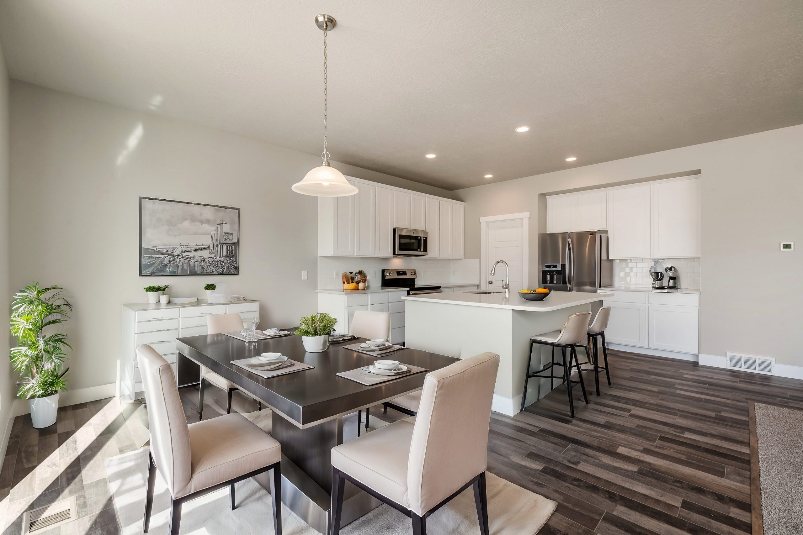 Modern kitchen and dining area found in many homes in Utah, featuring white cabinetry, stainless steel appliances, a dark wooden dining table for four, bar stools at the island, potted plants, and large windows flooding the space with natural light.