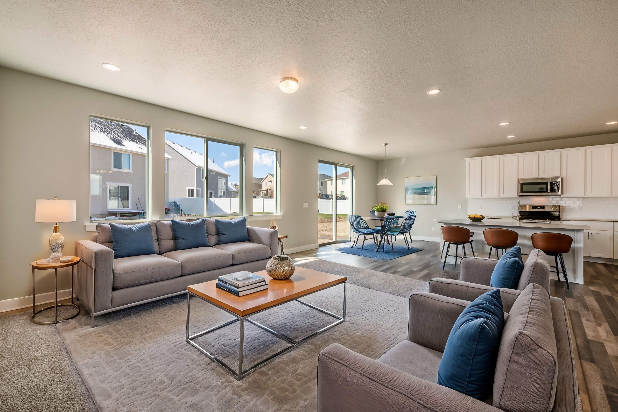 Bright, modern open-concept living room found in many homes in Utah, with gray sofas, blue accent pillows, a wooden coffee table, and a kitchen with white cabinets and barstools; large windows let in natural light.