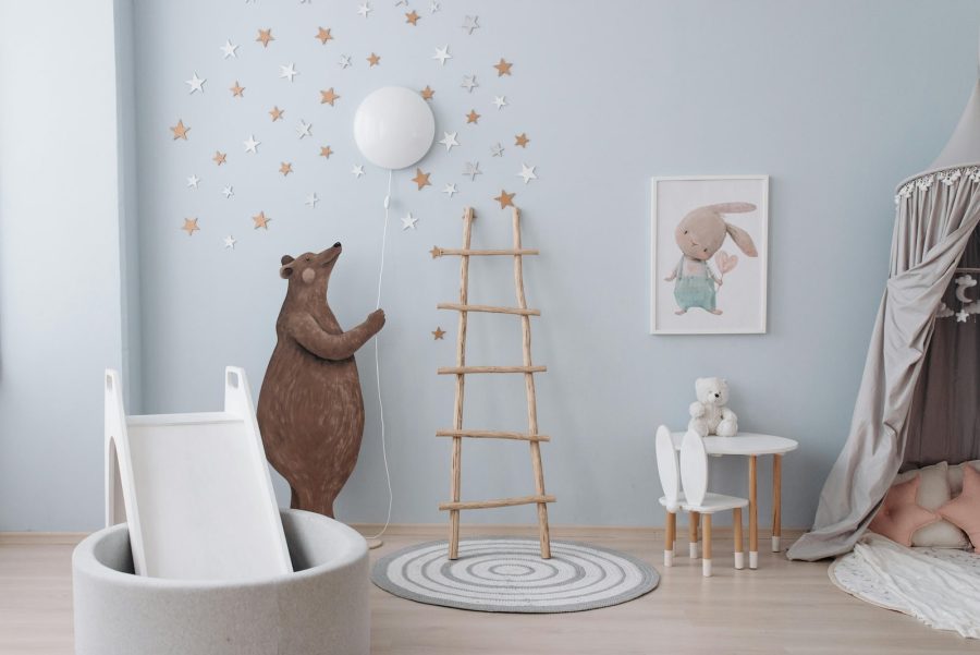 Whimsical children's playroom with a pastel blue wall, bear mural holding a balloon light, wooden ladder, round rug, small table and chair, indoor slide, and a cozy reading nook with star decorations and cushions.