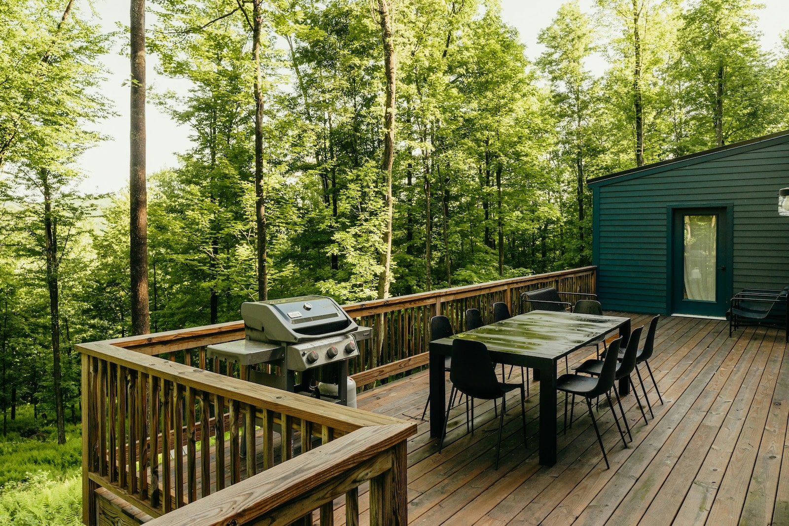 A spacious wooden deck surrounded by forest, featuring a black dining table with chairs and a stainless steel gas grill.