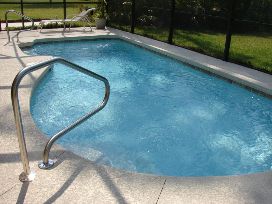 A small, clean backyard swimming pool with a curved stainless steel handrail and lounge chairs nearby, enclosed by a screen with green grass and trees outside.