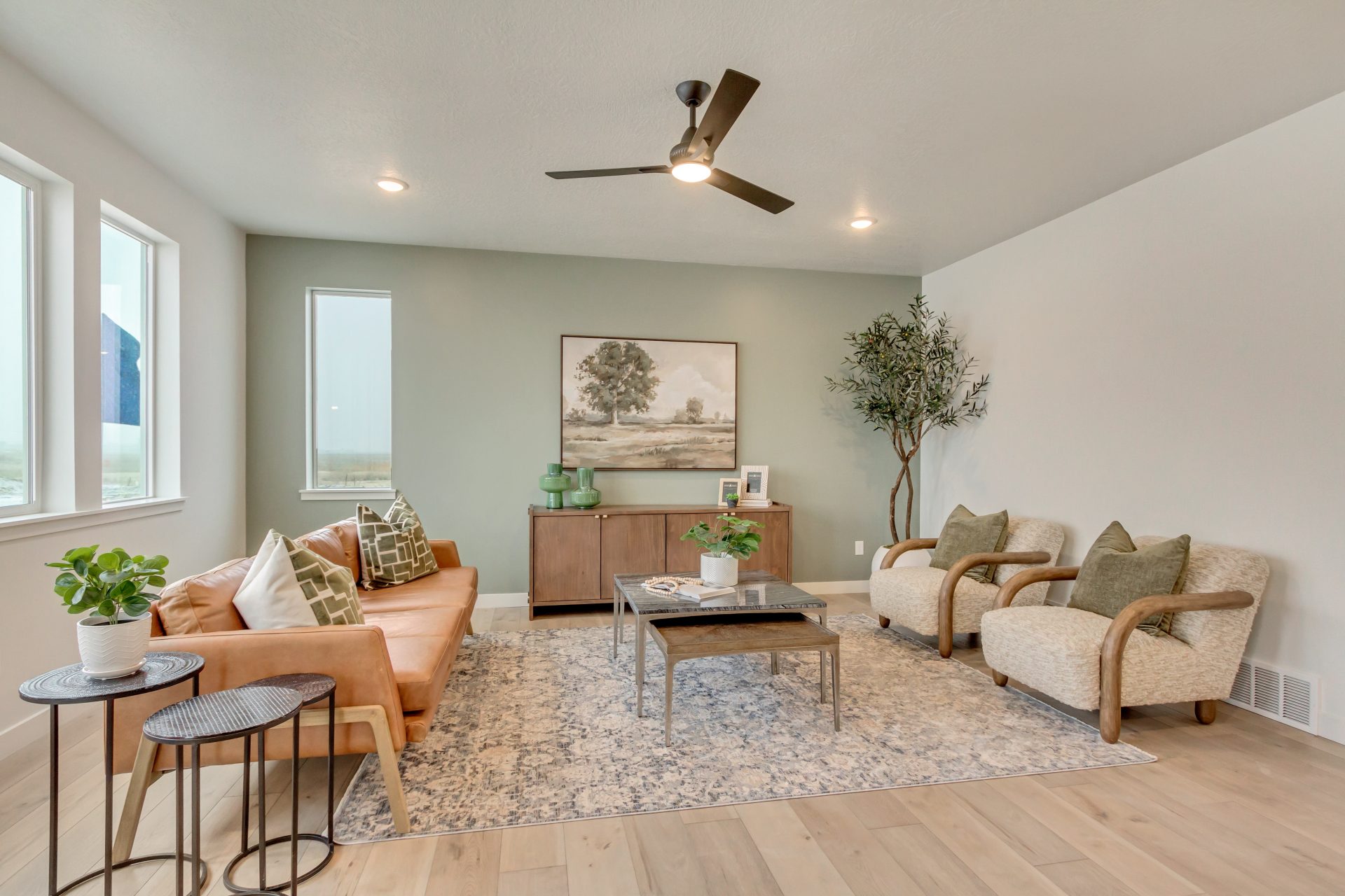 Modern living room with a tan leather sofa, two beige armchairs, a patterned rug, a wooden sideboard, green accent wall, large windows, indoor plants, and a ceiling fan with recessed lighting.