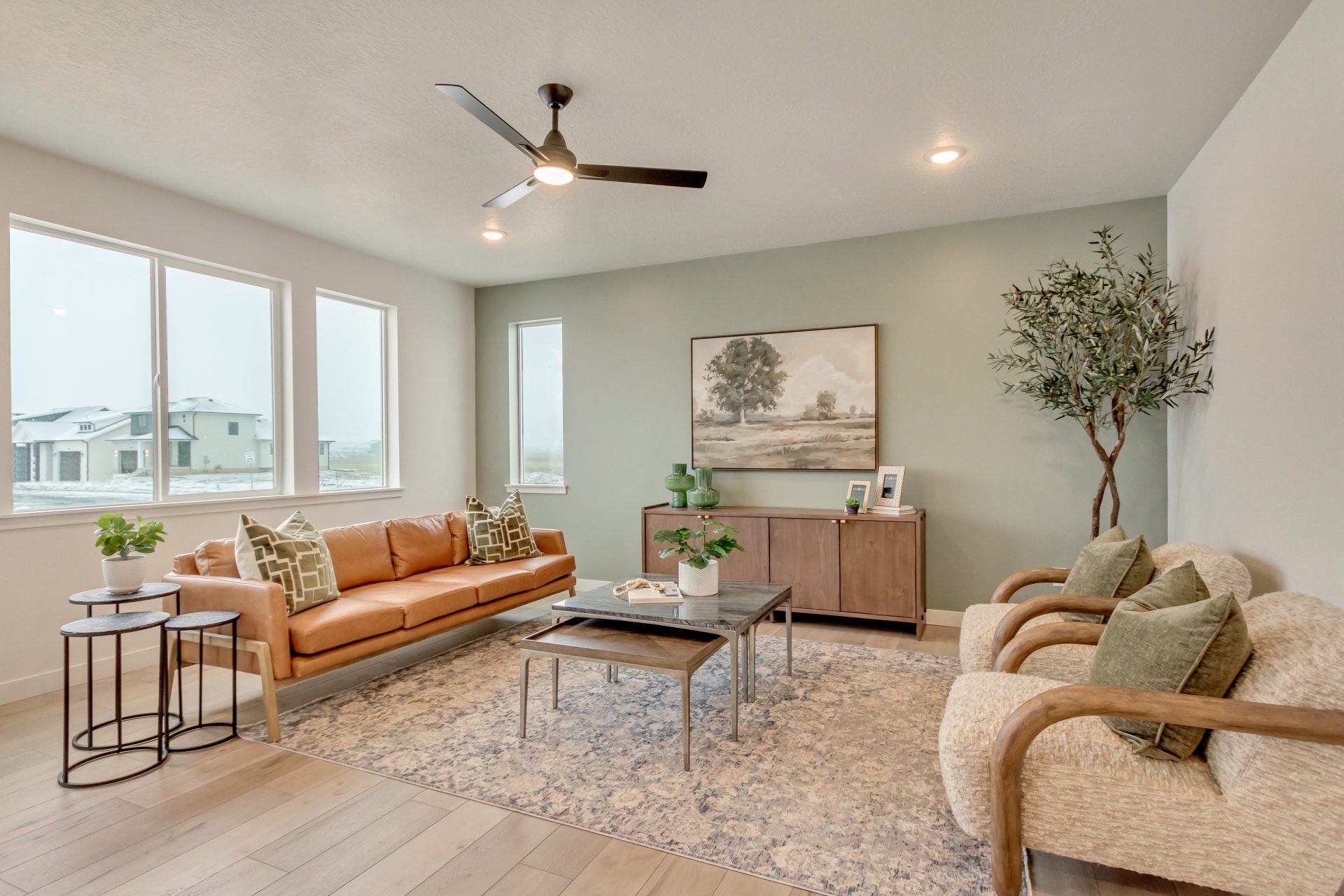Modern living room with a tan leather sofa, two beige chairs, a wooden coffee table, a side table, potted plants, a large wall art, and large windows letting in natural light.