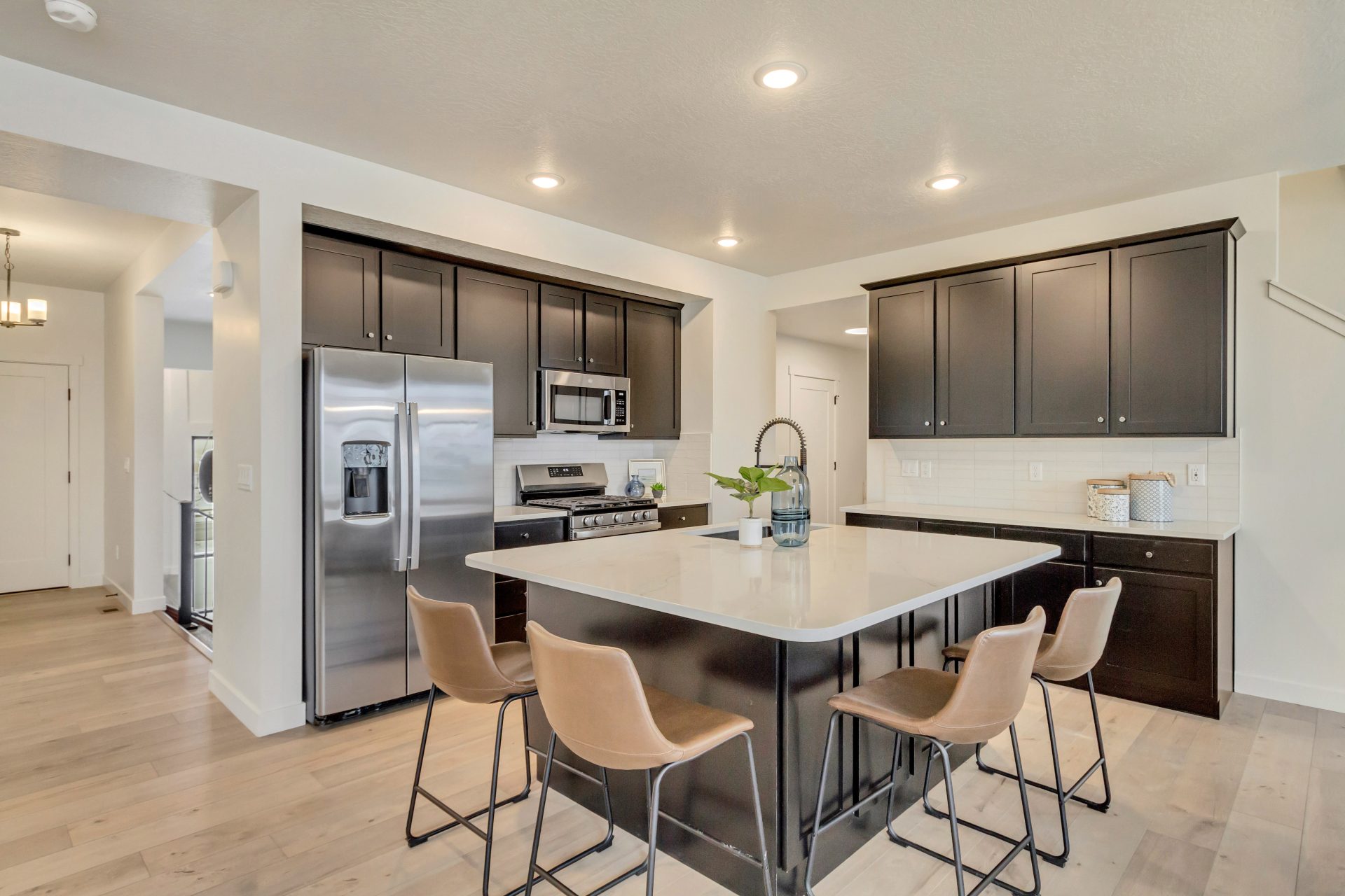Modern kitchen with dark cabinets, stainless steel appliances, a large island with a white countertop, four tan barstools, light wood flooring, and bright recessed lighting.