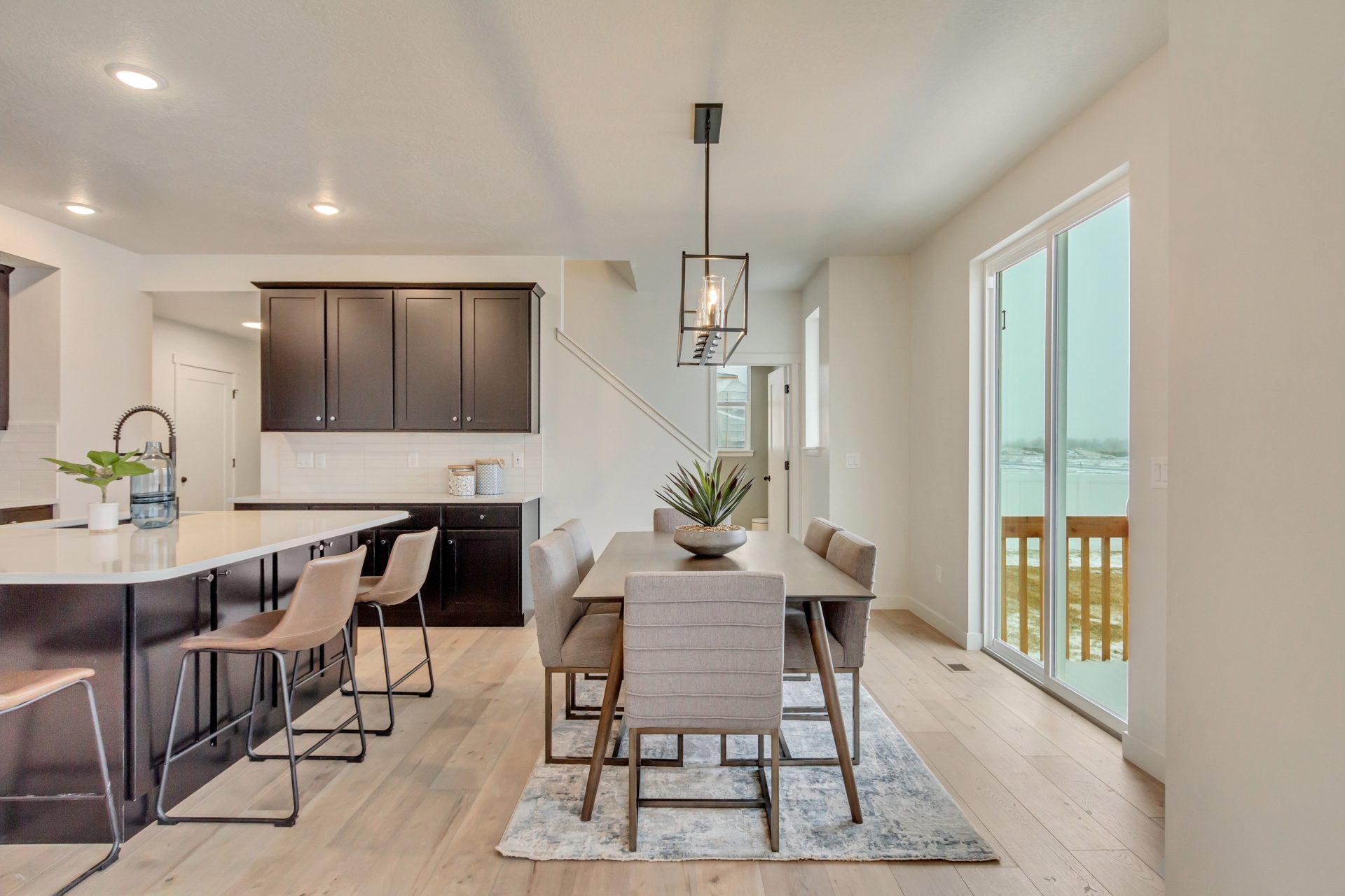 Modern dining and kitchen area with wooden floors, a rectangular dining table with six chairs, pendant lighting, kitchen island with stools, and large glass doors opening to a snowy outdoor deck.