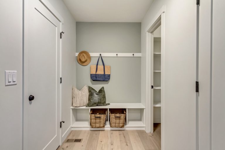 A tidy mudroom with light wood floors, a white bench, two wicker baskets, pillows, and wall hooks holding a hat and a tote bag. White walls and doors surround the space, with a closet visible on the right.