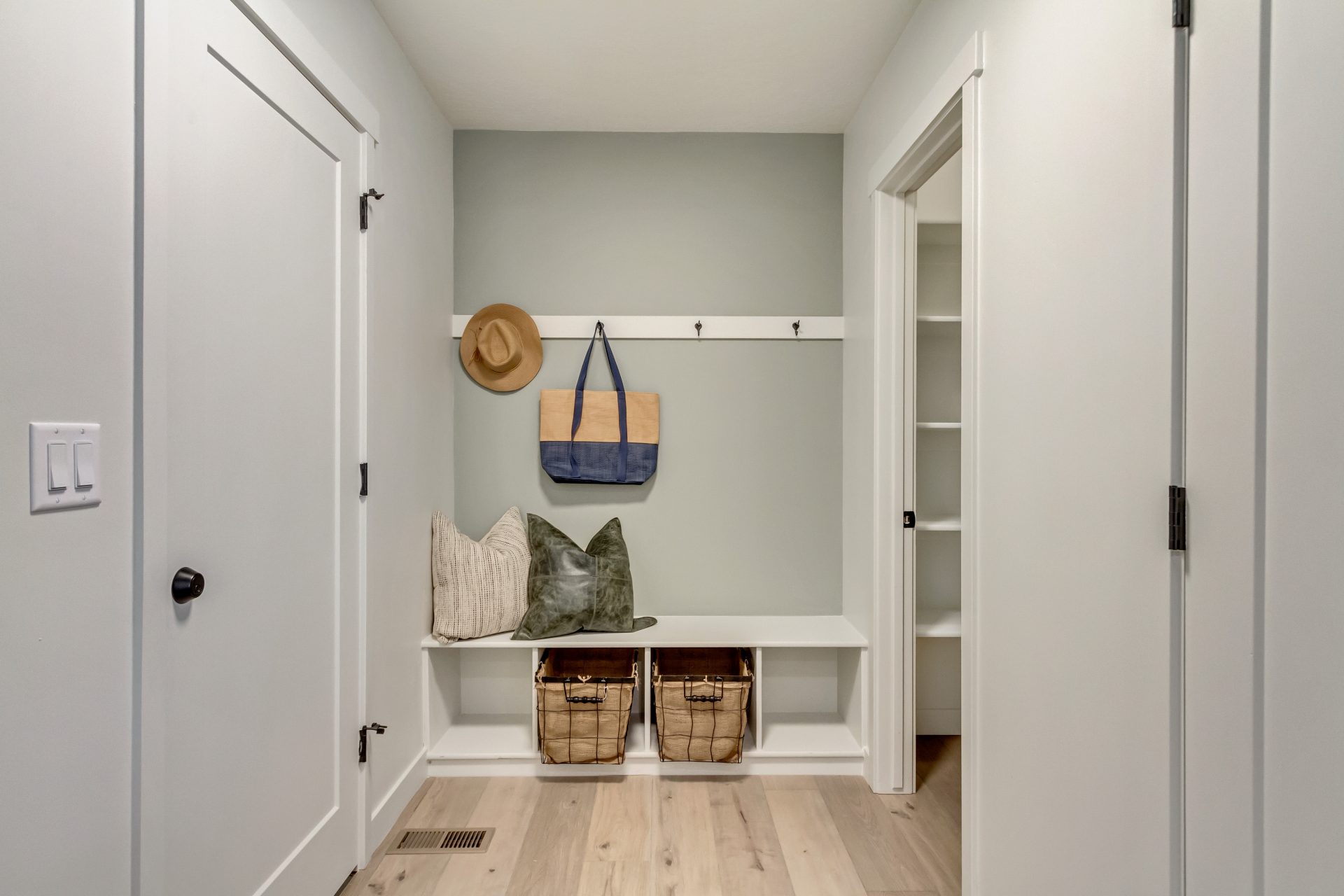 A tidy mudroom with light wood floors, a white bench, two wicker baskets, pillows, and wall hooks holding a hat and a tote bag. White walls and doors surround the space, with a closet visible on the right.