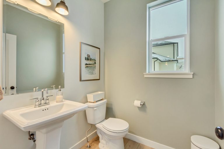 A modern bathroom with light gray walls, a white pedestal sink, a wall mirror with two lights above, a toilet with a tissue box, a toilet paper holder, wall art, and a window letting in natural light.
