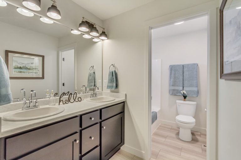 Modern bathroom with double sink vanity, two mirrors, and lights above. Towels hang on the wall and by the sinks. Through the open door, a toilet and a blue towel on a rack are visible. Neutral colors and clean decor.