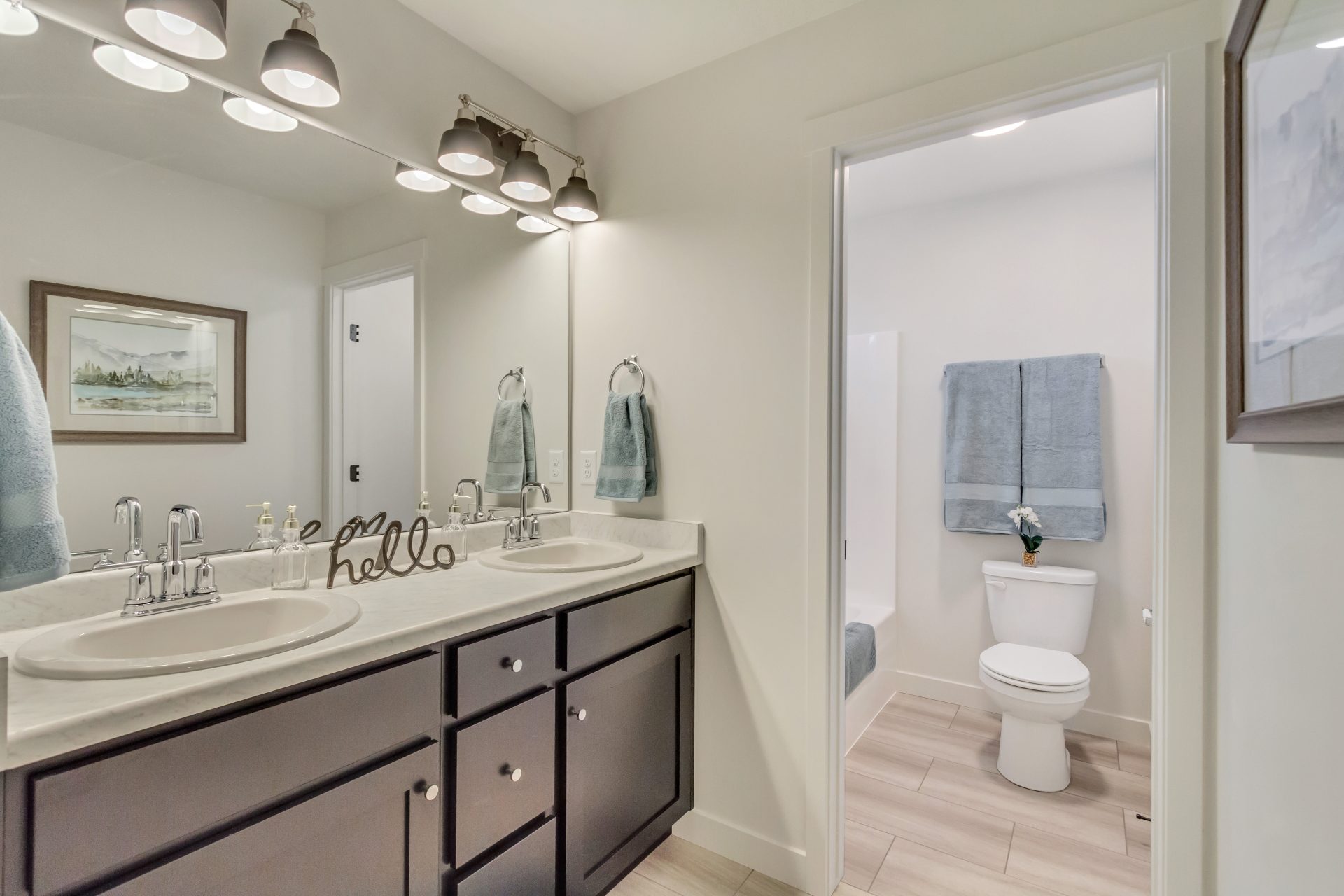 Modern bathroom with double sink vanity, two mirrors, and lights above. Towels hang on the wall and by the sinks. Through the open door, a toilet and a blue towel on a rack are visible. Neutral colors and clean decor.
