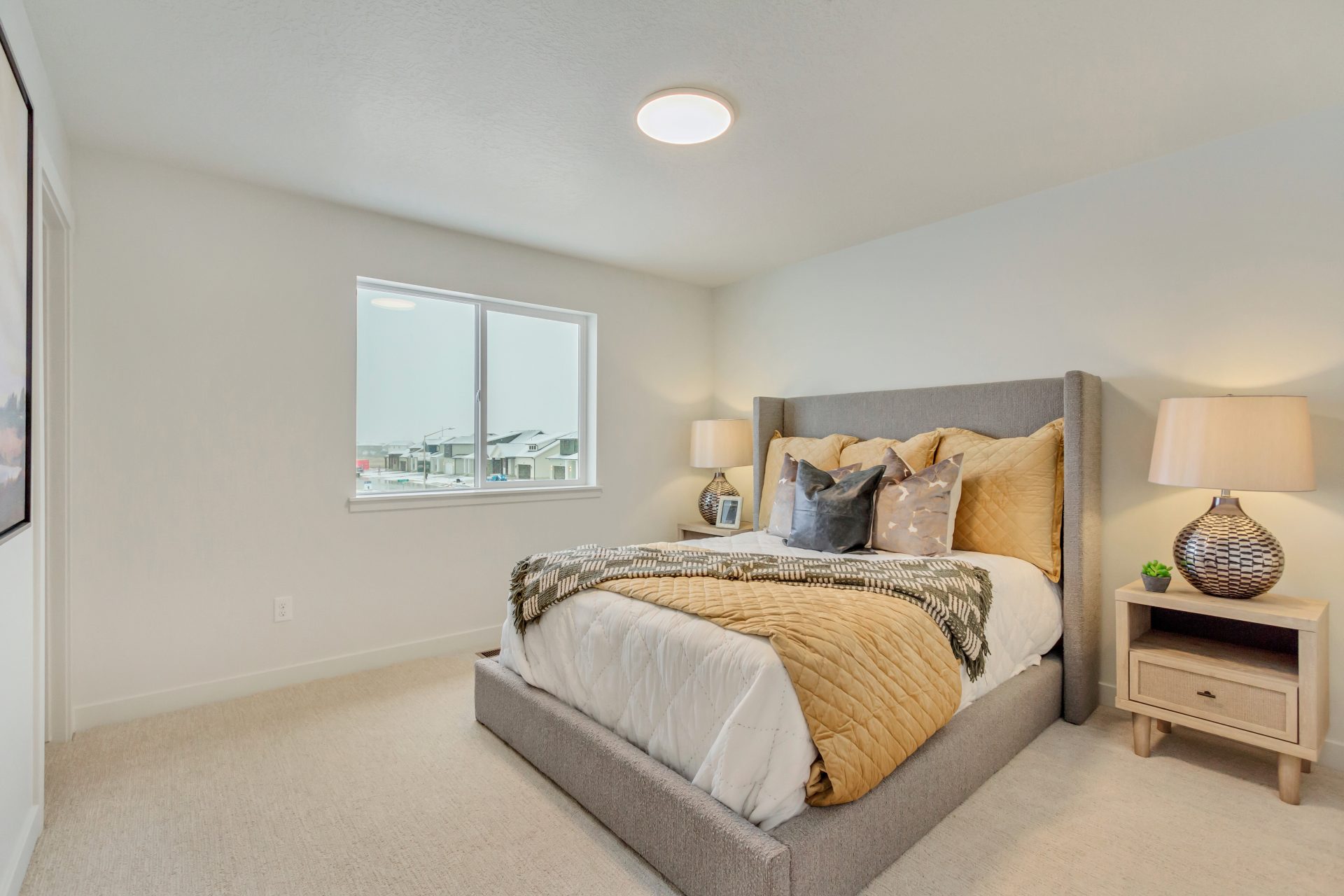 A cozy bedroom with a neatly made bed featuring gold and gray bedding, two bedside tables with lamps, a window letting in natural light, and neutral-colored walls and carpet.
