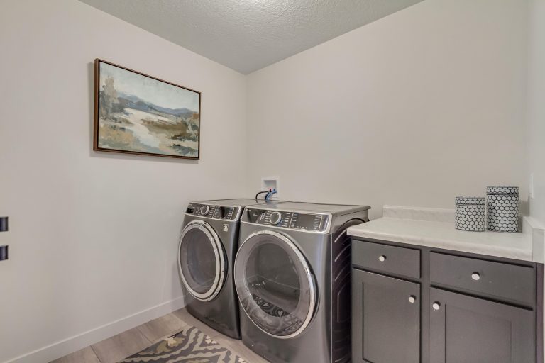 A modern laundry room with a front-loading washer and dryer, a patterned rug, a countertop with cabinets, two decorative containers, and a landscape painting on the wall.