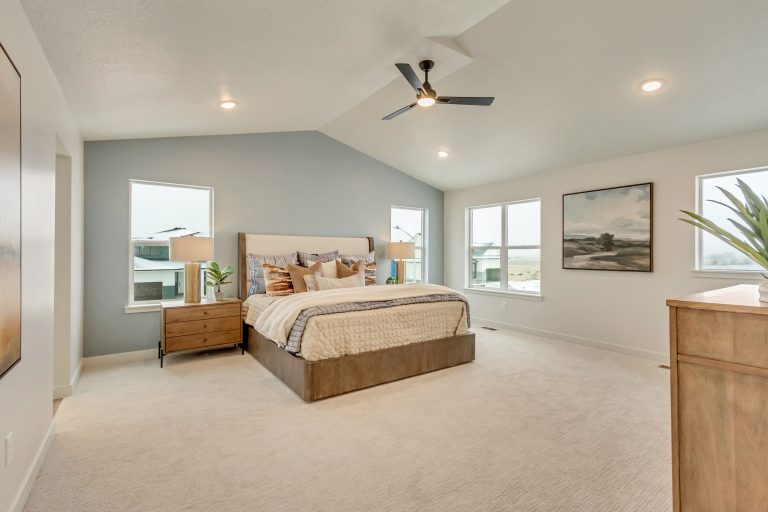 Spacious, modern bedroom with a large bed, neutral decor, nightstands and lamps on both sides, artwork on the wall, dresser with a plant, beige carpet, and two windows letting in natural light.