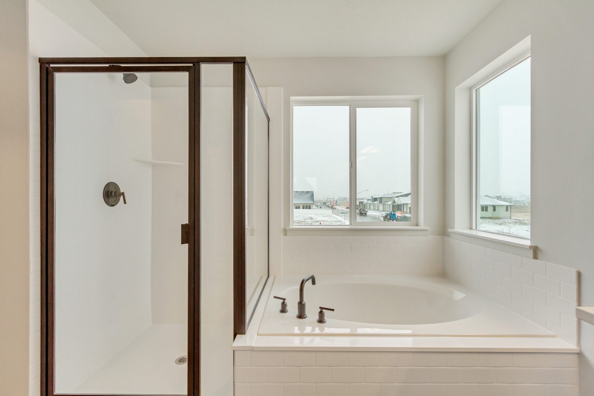 A modern bathroom with a glass-enclosed shower next to a large soaking bathtub, both surrounded by white tile, with two windows letting in natural light and a view of houses outside.