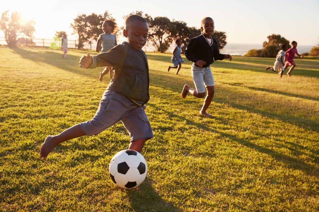 Children playing soccer in a grassy park at sunset, with one child kicking a soccer ball while others run and laugh in the background.
