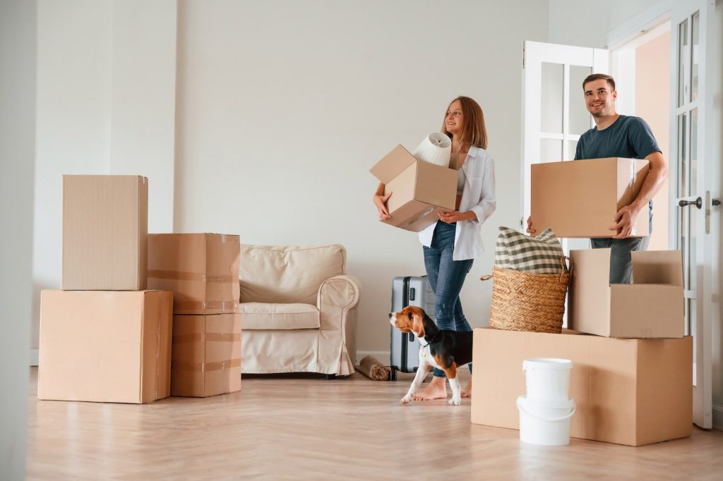 A smiling couple carries moving boxes into a bright, unfurnished living room. A dog stands near their feet, and several boxes, a suitcase, and household items are scattered around the wooden floor.