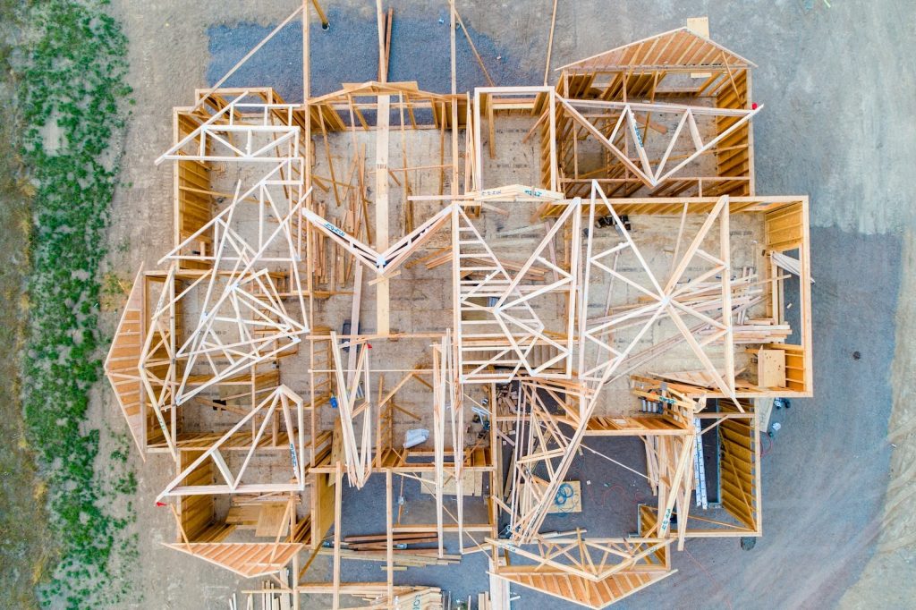 Aerial view of a house under construction, showing exposed wooden framing, trusses, and beams forming the structure of the building on a dirt lot.
