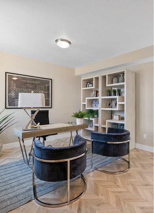 A modern home office with a wooden desk, two dark velvet chairs with metal frames, a gray rug, bookshelf with decor and books, a framed print on the wall, and a potted plant in the corner.