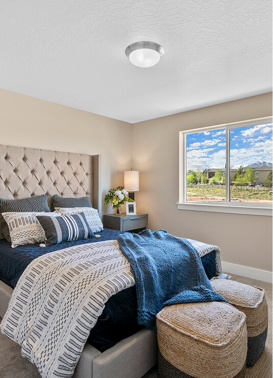 A modern bedroom with a tufted headboard, neatly made bed with blue and gray bedding, two woven ottomans at the foot, a nightstand with a lamp and flowers, and a window showing a bright sky and greenery outside.