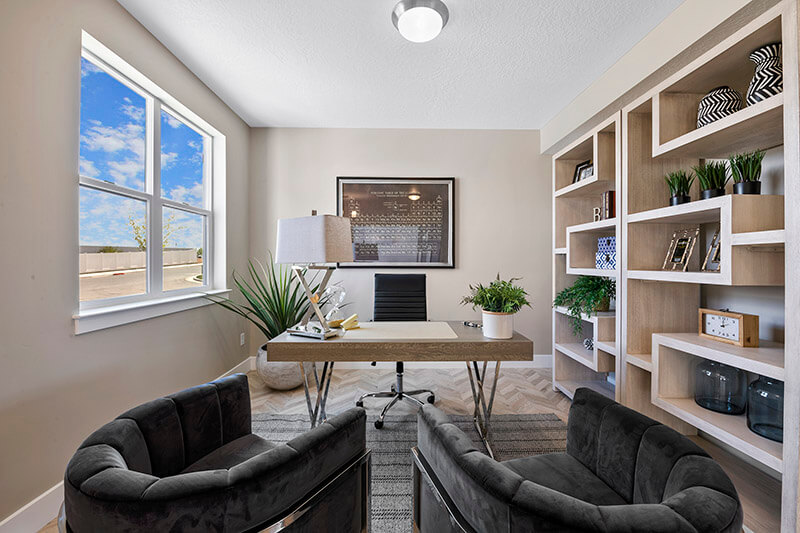 A modern home office with a wooden desk, black chair, and two gray armchairs. Built-in shelves with books and plants line one wall, and a large window lets in natural light. A potted plant and lamp sit on the desk.