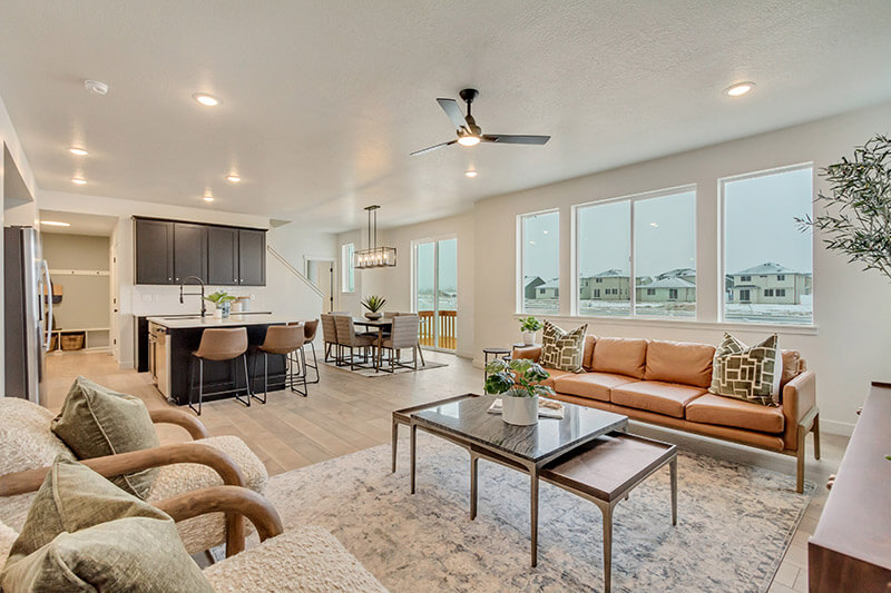 Modern open-concept living room and kitchen with large windows, tan sofa, armchairs, a coffee table, bar stools at a dark island, dining area, and neutral decor, with snowy houses visible outside.