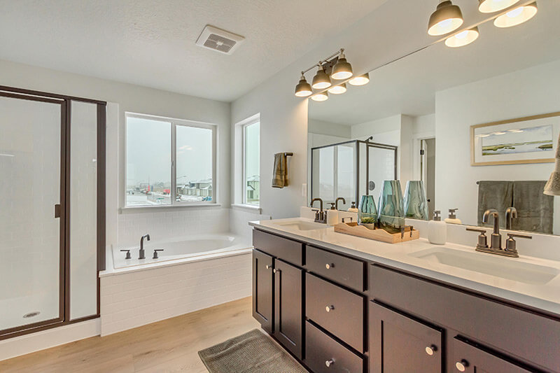 Modern bathroom with a glass shower, large bathtub near two windows, double sinks with dark cabinets, a large mirror, and decorative vases on the counter. Neutral tones and bright natural light fill the room.