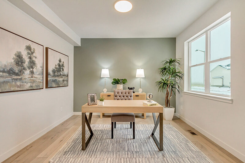 A modern home office with a wooden desk, tufted chair, two lamps on a sideboard, wall art, a potted plant, and a large window letting in natural light. The decor is minimalist and neutral-toned.