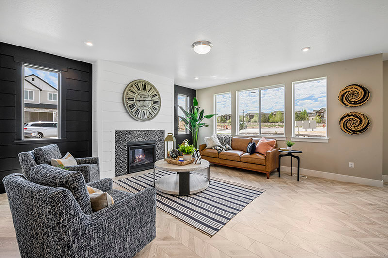 Modern living room with a fireplace, large wall clock, patterned armchairs, tan sofa, striped rug, round coffee table, wall art, potted plant, and three large windows letting in natural light.