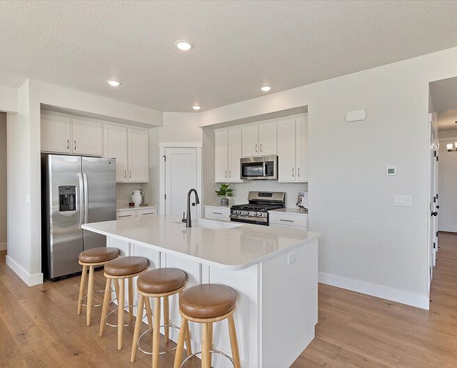 Modern kitchen with white cabinets, stainless steel appliances, a center island with a white countertop, and four wooden bar stools on a light hardwood floor. Recessed lighting brightens the space.