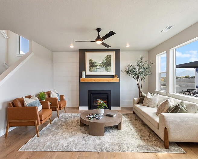Bright living room with tan armchairs, a cream sofa, a round wooden coffee table, fireplace with wooden mantel, wall art, potted plant, and large windows letting in natural light. Neutral tones throughout.