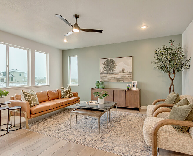 Modern living room with a tan sofa, two armchairs, coffee table, and side tables on a patterned rug. Large windows, green accent wall with landscape art, potted plant, and ceiling fan complete the space.