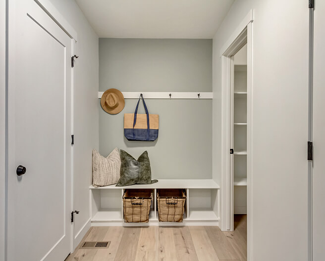 A mudroom with light wood floors, a built-in white bench, two storage baskets underneath, three pillows, a straw hat, a blue tote bag on hooks, and white walls with a gray accent wall.