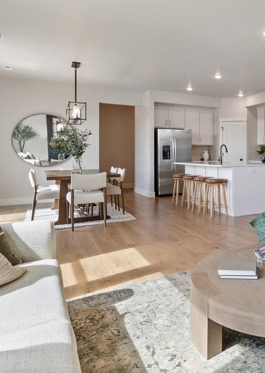 A modern open-concept kitchen and dining area with light wood floors, a round dining table, a large mirror on the wall, a stainless steel refrigerator, a kitchen island with bar stools, and a cozy living area with a beige sofa.