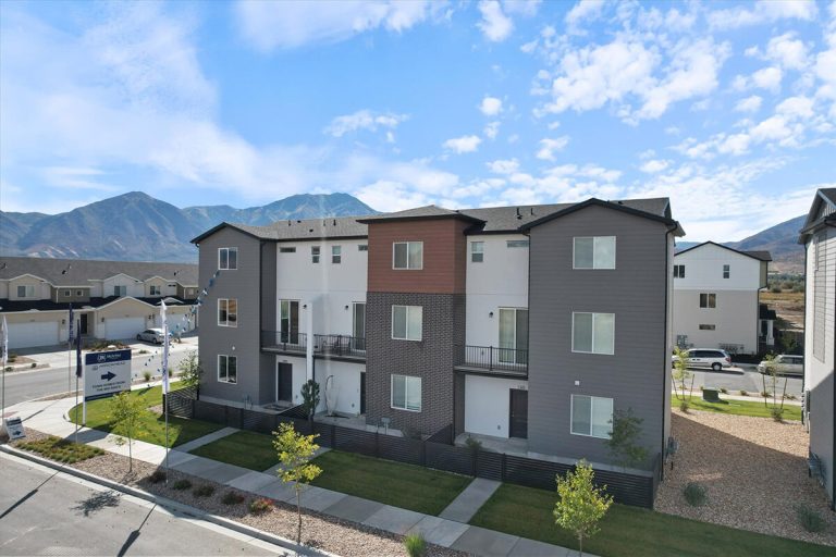 A modern three-story apartment building with balconies, large windows, and a landscaped lawn, set against a backdrop of mountains under a partly cloudy sky.