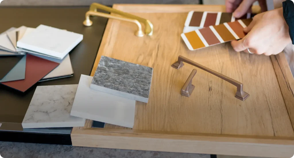 Hands compare paint swatches beside brass and bronze cabinet pulls, marble and stone countertop samples while picking home finishes.