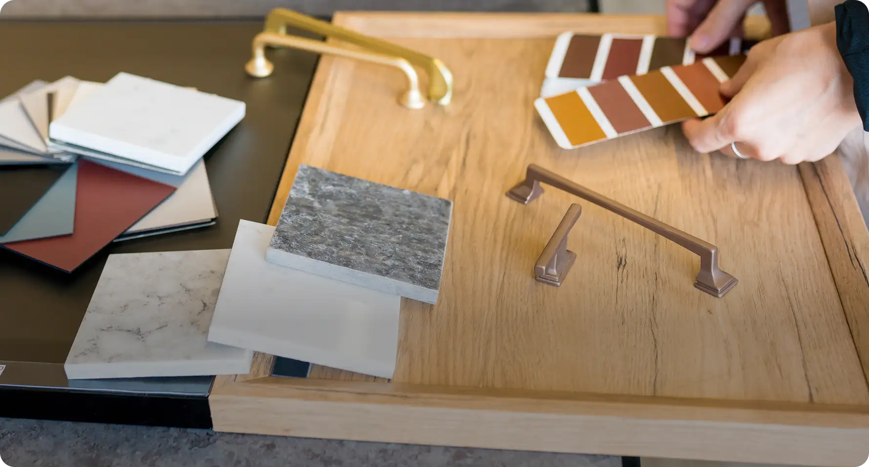 Hands compare paint swatches beside brass and bronze cabinet pulls, marble and stone countertop samples while picking home finishes.
